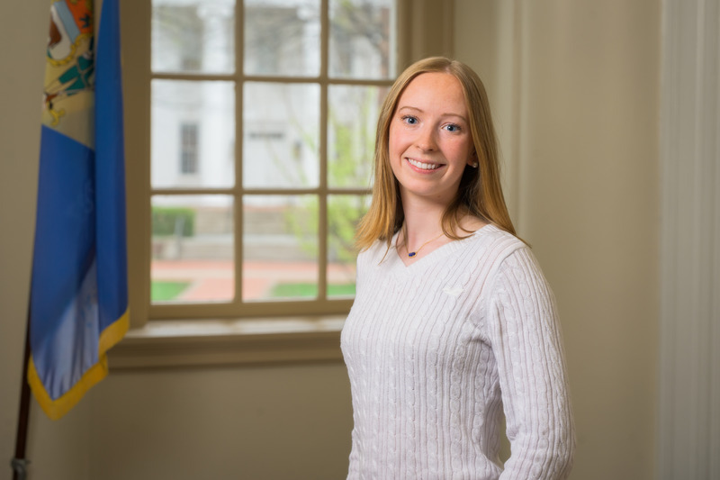A college student stands in front of a sunny window and blue flag.