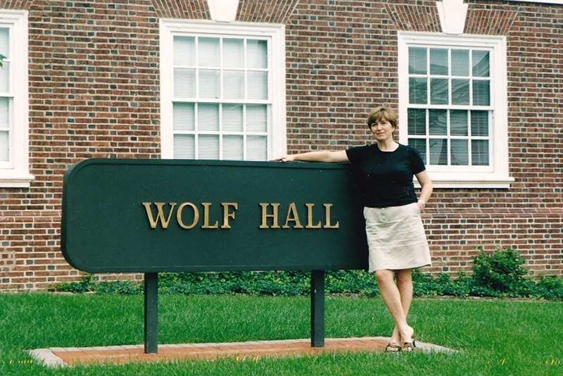 A college student standing next to a sign for Wolf Hall, located in front of a red brick academic building