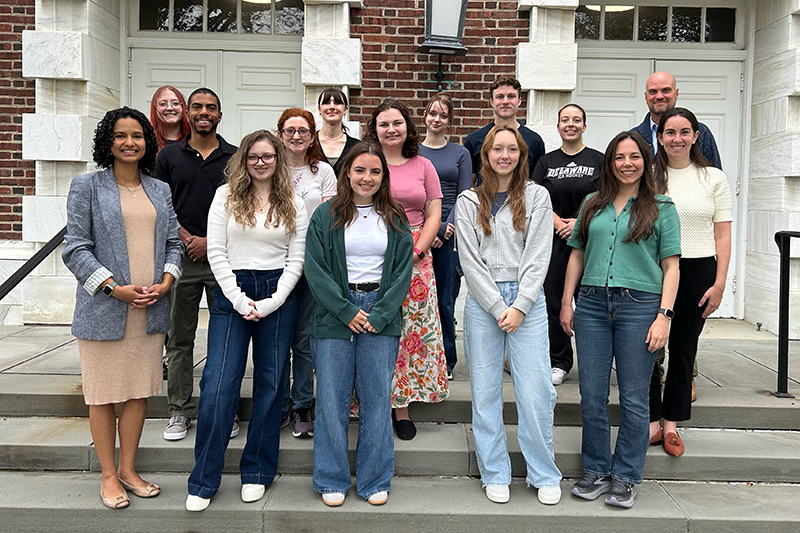 A group of 15 students and faculty stand on the steps in front of a red brick academic building