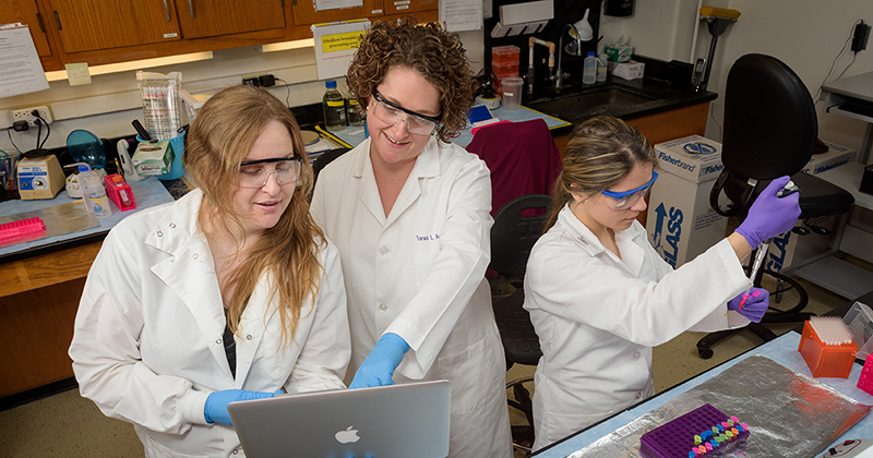 A professor and two female graduate students, all wearing white lab coats, latex gloves, and safety classes, review lab results recorded in a notebook while standing inside a laboratory.