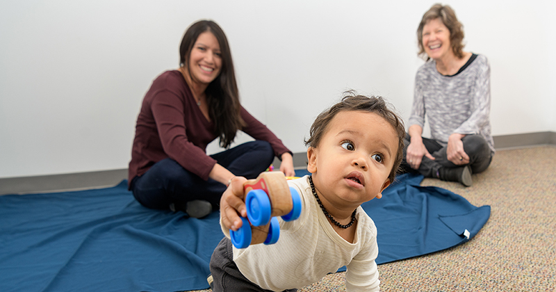 A young toddler crawls in the foreground, holding a toy, while the mother and college researcher looks on.