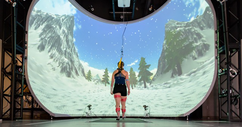 Image of the back of a woman walking in from of a projection of a winter scene with two camera pointing at her. She wears sensors and a harness.
