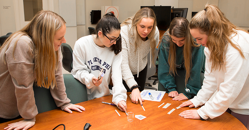 A group of college students inside a classroom work together around a table to solve a series of puzzles. They 