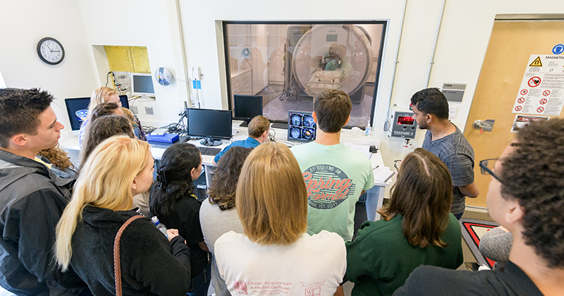College students visit a functional magnetic resonance imaging lab to learn about brain imaging. They observe the professor, who uses desktop computer to operate the scanner, visible through a window in the adjacent room.