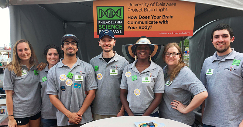 A group of University of Delaware graduate students, all wearing gray polo shirts, stand together inside their booth at the Philadelphia Science Festival, in front of a sign that reads: University of Delaware Project Brain Light: How Does Your Brain Communicate with Your Body?