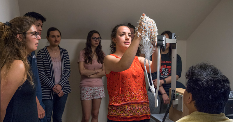 A group of neuroscience students observe their instructor as she prepares to place a cap filled with electrodes on a man's head for a brain-mapping procedure.