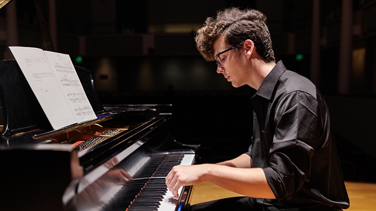 Music composition student performs his piece on a black grand piano on a stage.