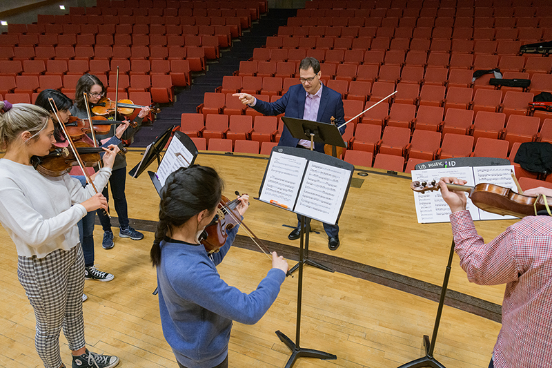 Students stand in circle on a theatre stage, playing violins and learning about music