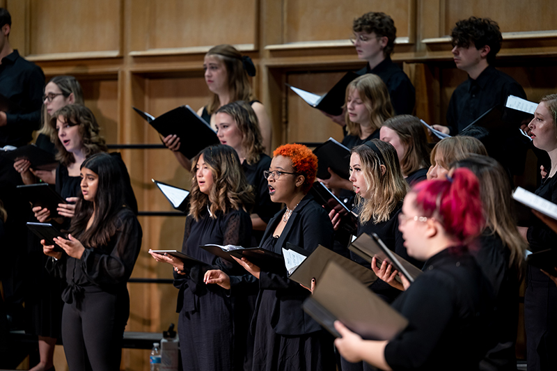 A choir of college students perform on stage while holding their music books.
