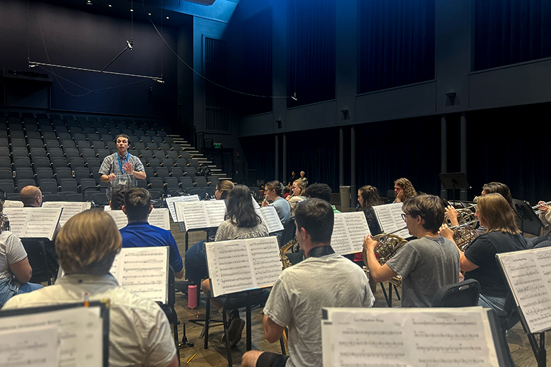 Student band inside an empty theater plays instruments, using sheet music on stands, with a conductor instructing them in the background.
