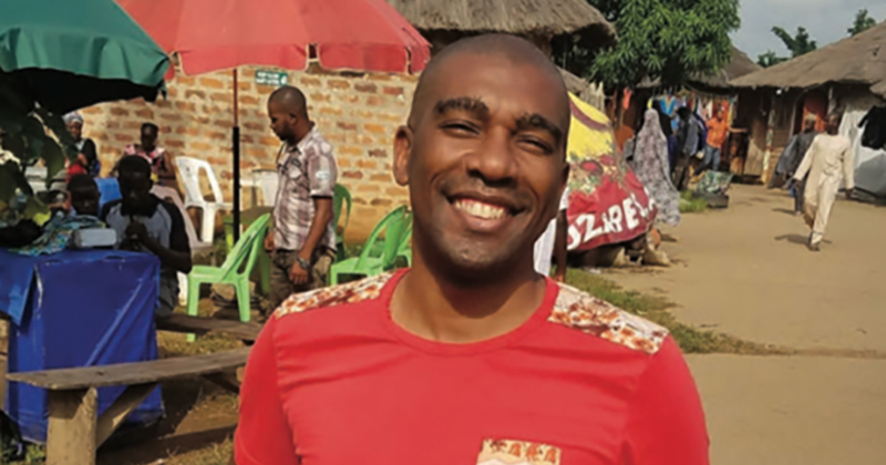 Jullion Cooper, dressed in a red t-shirt, stands in the middle of a Haitian village with buildings, people and colorful umbrellas in the background.