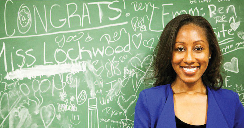 Woman in classroom stands in front of a green blackboard filled with congratulatory messages.
