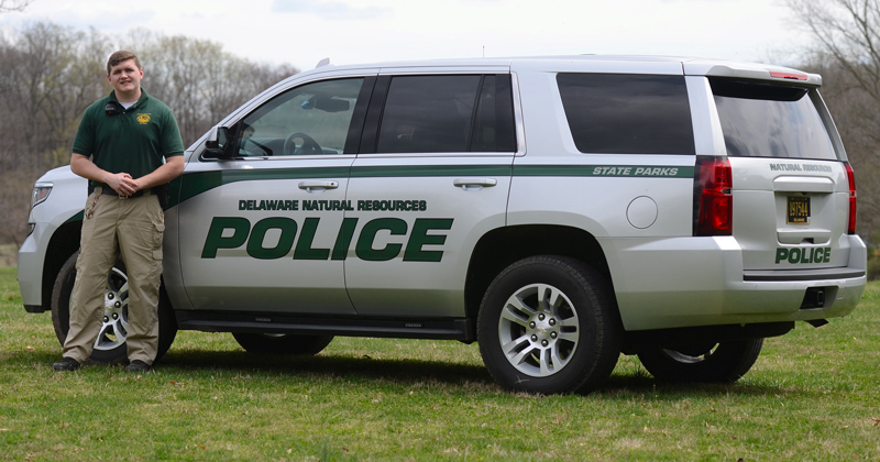 Brandon Pellegrini, a senior criminal justice major at UD, wears a green polo shirt and stands next to the white Delaware Natural Resources Police SUV, which sits on grass in at White Clay Creek State Park.