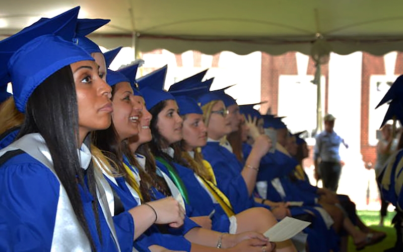 A row of University of Delaware sociology students dressed in blue caps and gowns and seated in a tent on The Green listen to remarks during their convocation ceremony.