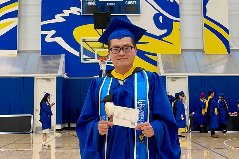 Tong Li in front of a Blue Hen painting while wearing a graduation gown and cap.