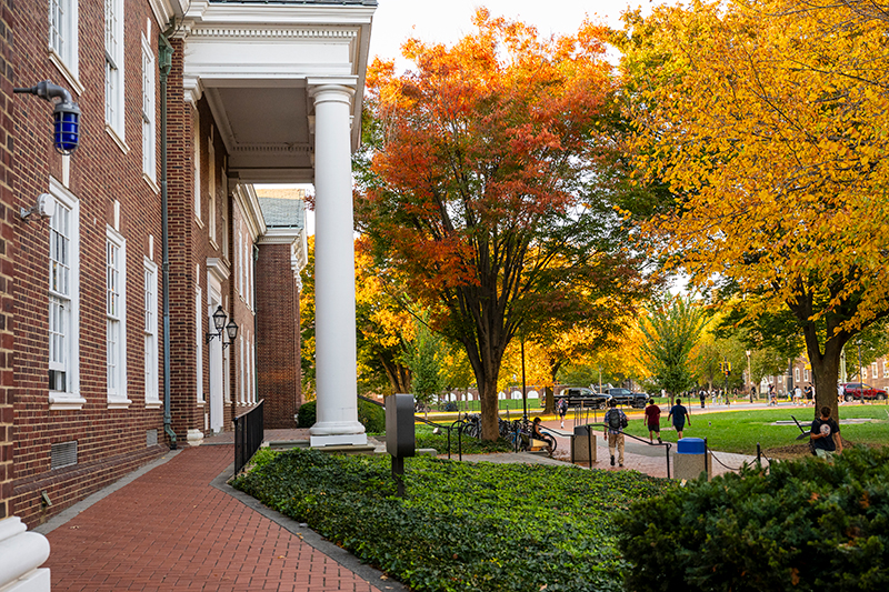 Gore Hall at the University of Delaware Side view of a historic red brick building with white columns and a brick walkway lined with trees with colorful fall leaves.