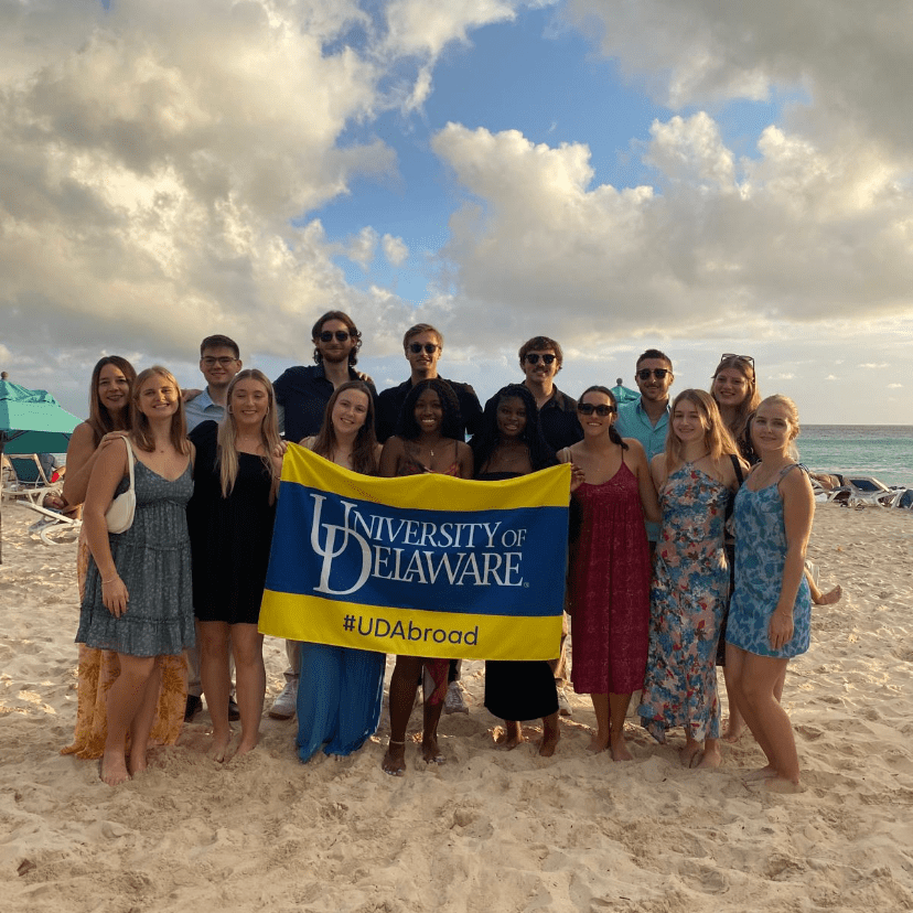A group of college students on a sandy ocean beach hold a University of Delaware banner.