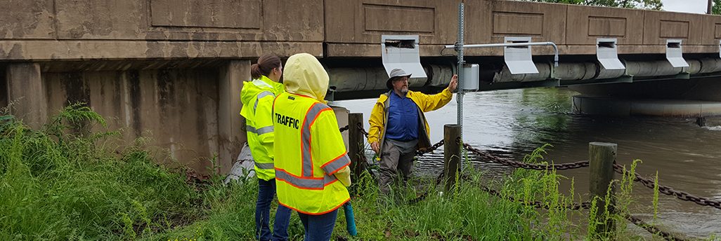 Standing by sensing equipment next to a creek below a bridge, David Huntley of the Center for Environmental Monitoring and Analysis explains low-cost flood sensors he and his team built in a pilot for the Delaware Department of Transportation.