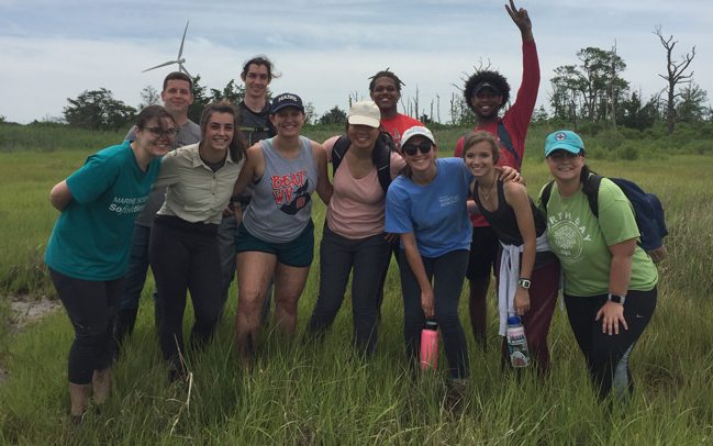 Marine Science students posing for a group photo in the Great Marsh, wind turbine showing in background
