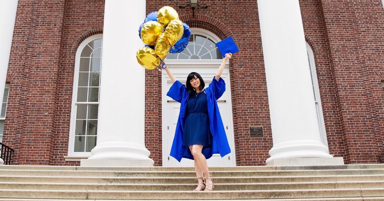Chelsea Lee wears a blue graduation robe and holds balloons and her grad cap in front of Memorial Hall