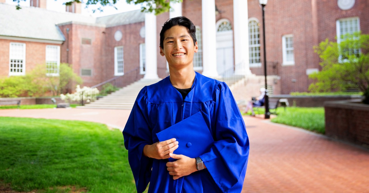 Parker Chan wears a blue graduation robe and holds his cap in front of Memorial Hall