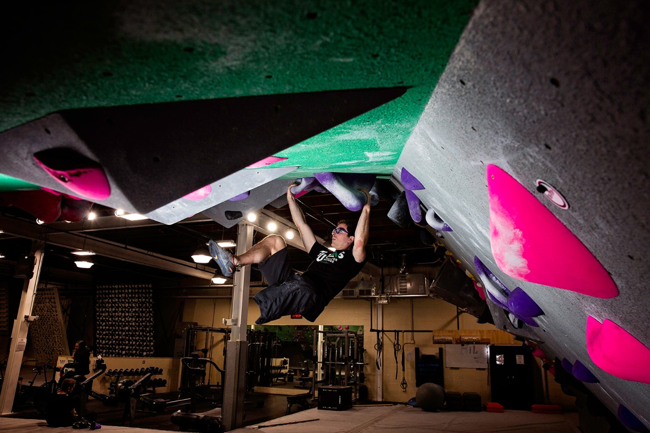 Travis Pollen, an amputee wearing a black T-shirt and shorts, hangs from a colorful angled rock wall