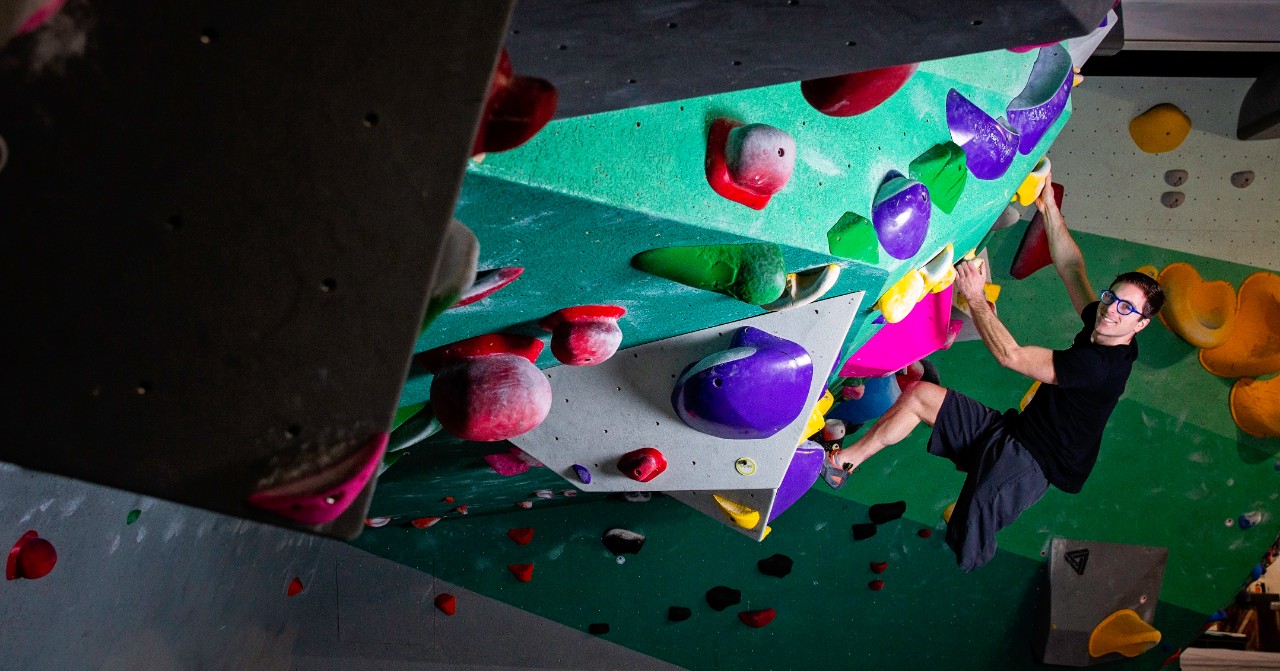Travis Pollen, an amputee wearing a black T-shirt and shorts, hangs from a colorful angled rock wall
