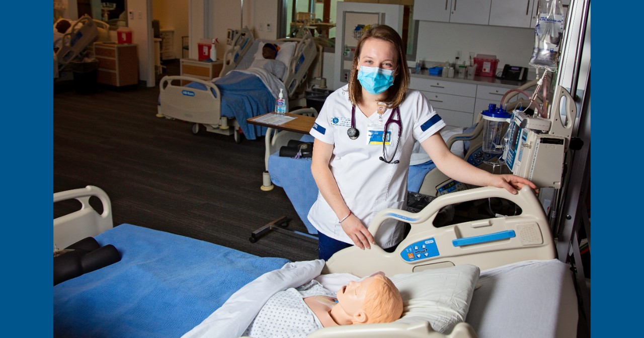 Shannon Gibbons wears UD nursing scrubs and stands next to a classroom hospital bed with a manikin patient laying in it