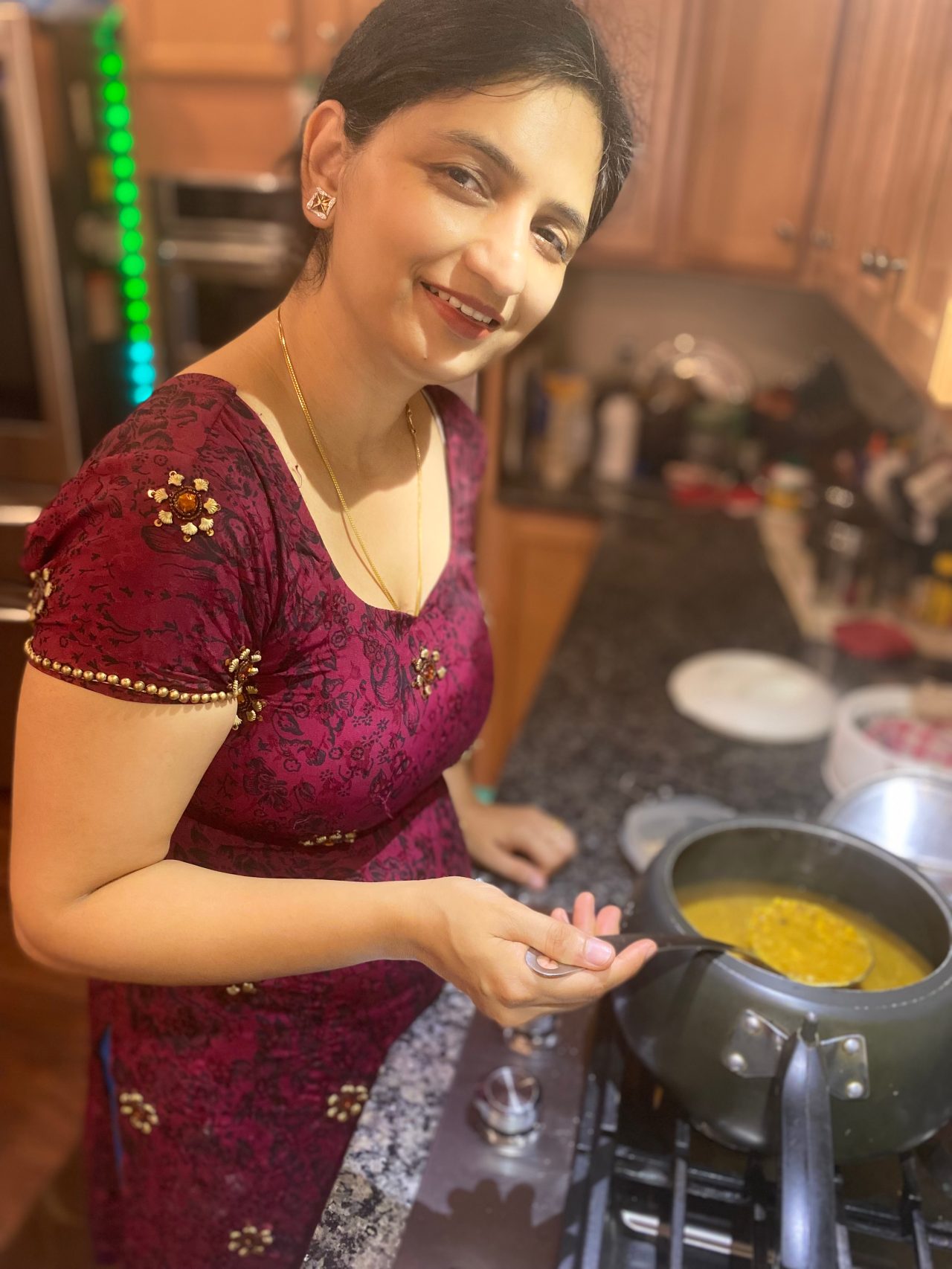 Mona Batish, wearing a maroon top, leans over a pot of yellow soup on the stove