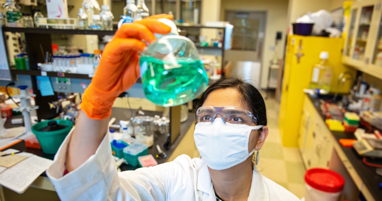 CHS-Humans-Mona-Batish-042721 Mona Batish, wearing a white lab coat, holds up a vial filled with green liquid in the middle of a busy wet lab