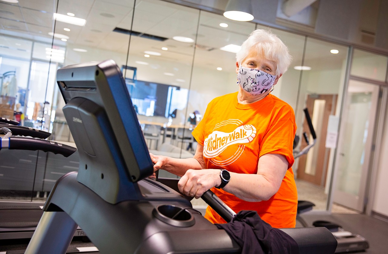 Bev LaRock wears an orange T-shirt and face mask while working out on a treadmill