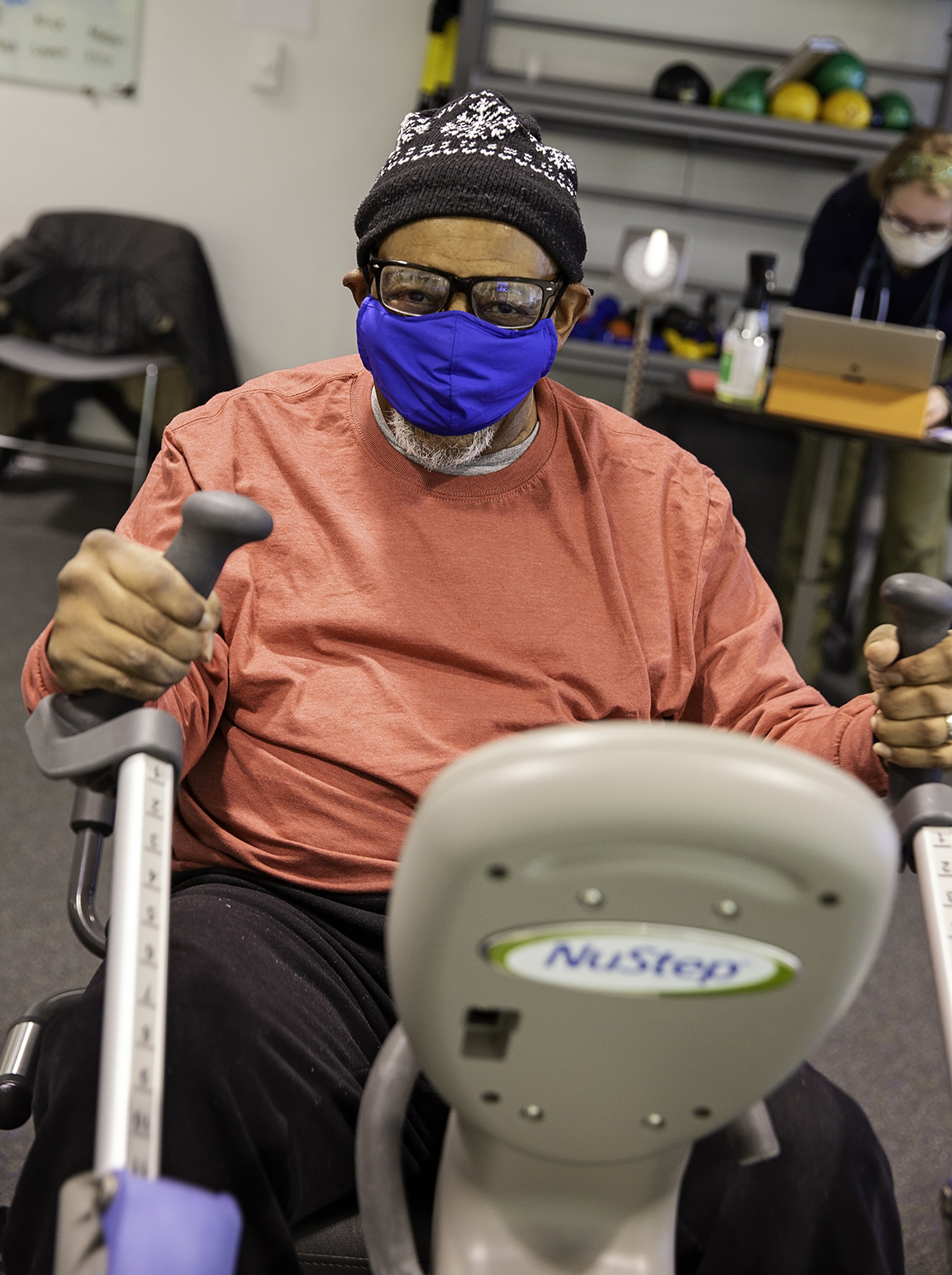 Joe Cratic, wearing a salmon long sleeve shirt, blue face mask and glasses, works out on an exercise bike