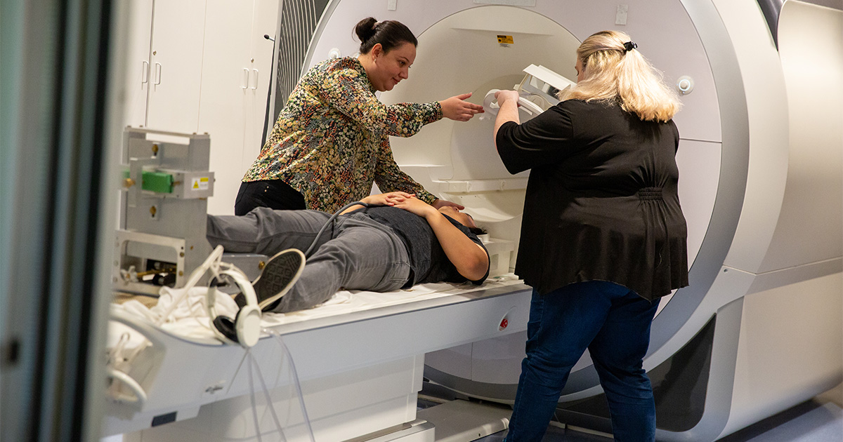 A man lies in a MRI scanner with a specialized foot attachment that helps keep the head still to get useful MRI scans in patients with Parkinson's disease. In this photo, Assistant Kinesiology Professor Roxana Burciu helps ready the device along with research assistant Abigail Bower.