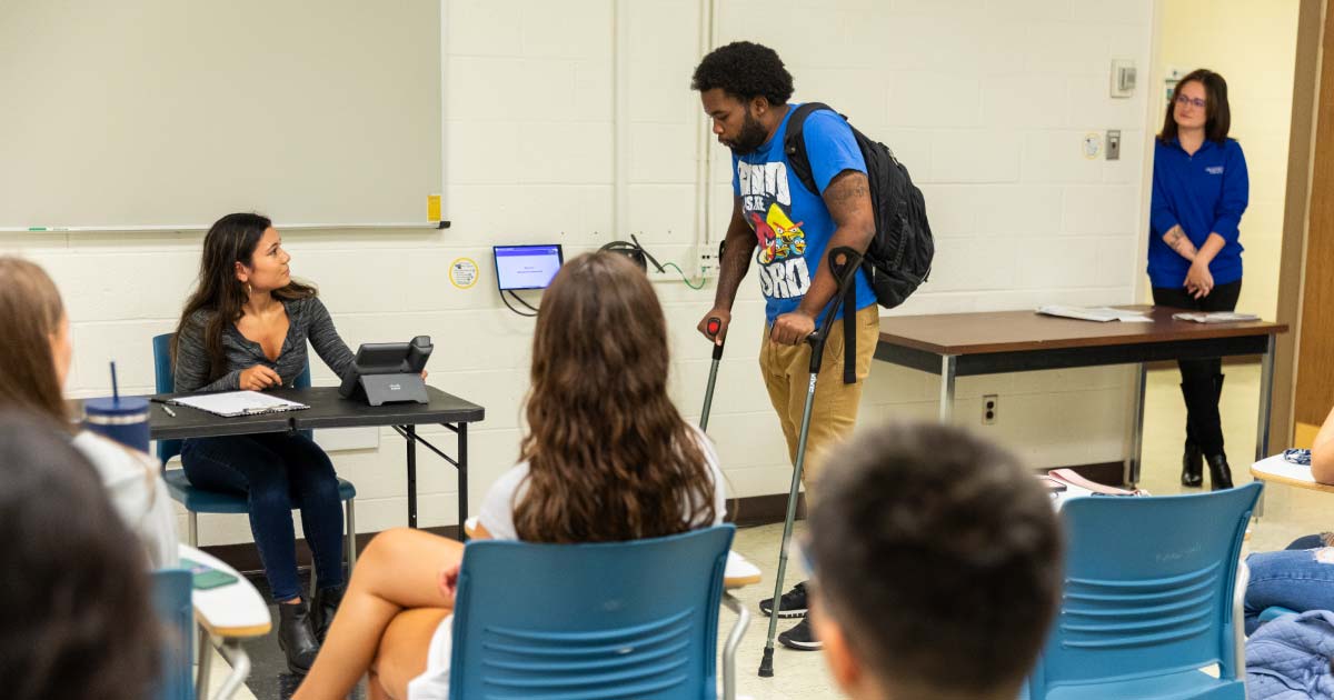 A man with crutches walks to meet a student acting as nurse at a desk as part of a presentation for new students