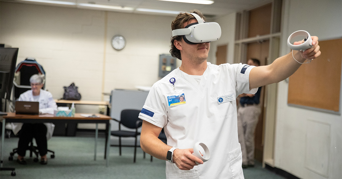 School of Nursing student Luke Stuchlik wears his nursing uniform and an Oculus headset while extending one arm out using the controller as he's transported from a classroom in McDowell Hall into a hospital setting as part of virtual reality simulation training.
