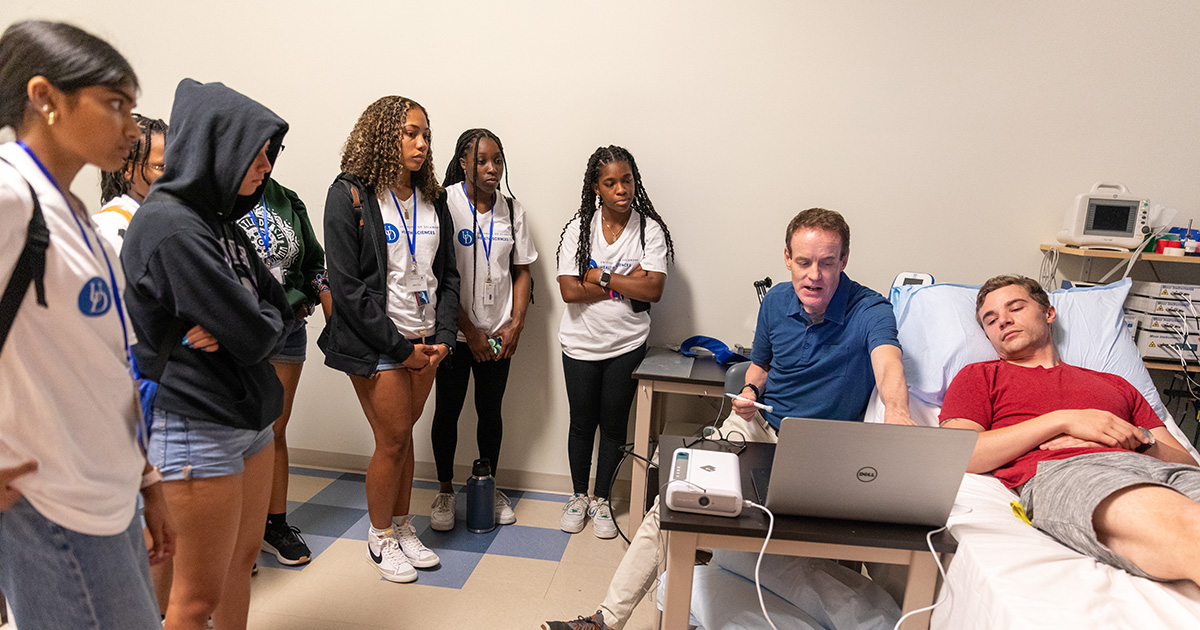 Dave Edwards, new chair of the Department of Kinesiology and Applied Physiology, points at data on a computer screen while a patient or subject lies down and students from the College of Health Sciences Summer Camp learn about the impacts of excess sodium on cardiovascular function.