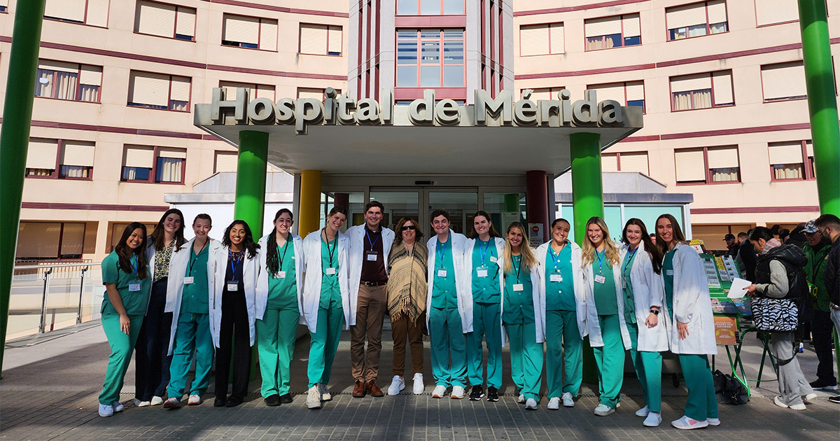 College of Health Sciences and College of Arts and Sciences students, wearing green scrubs and white lab coats, pose with Medical and Molecular Sciences associate professor Virginia Hughes outside Hospital de Mérida in Spain, where they shadowed physicians over Winter Session.