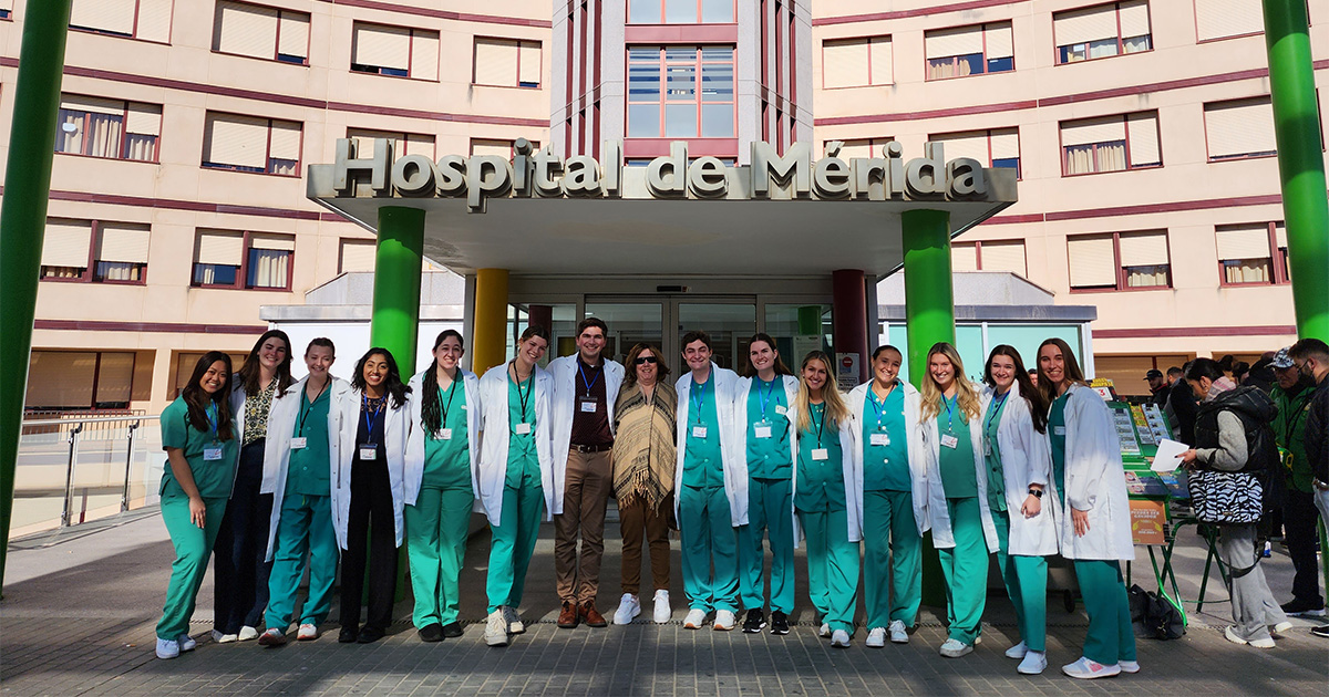 College of Health Sciences and College of Arts and Sciences students, wearing green scrubs and white lab coats, pose with Medical and Molecular Sciences associate professor Virginia Hughes outside Hospital de Mérida in Spain, where they shadowed physicians over Winter Session.