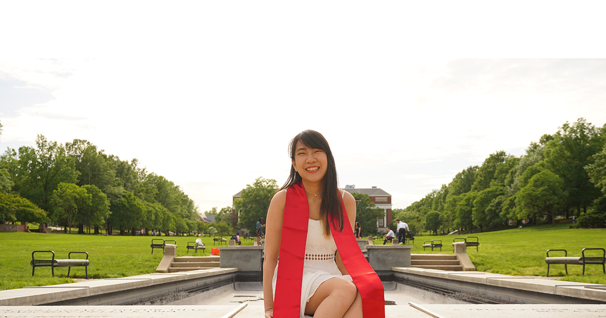 UD alumna Christina Tao poses for a photo while sitting outdoors with lots of green space behind her. 