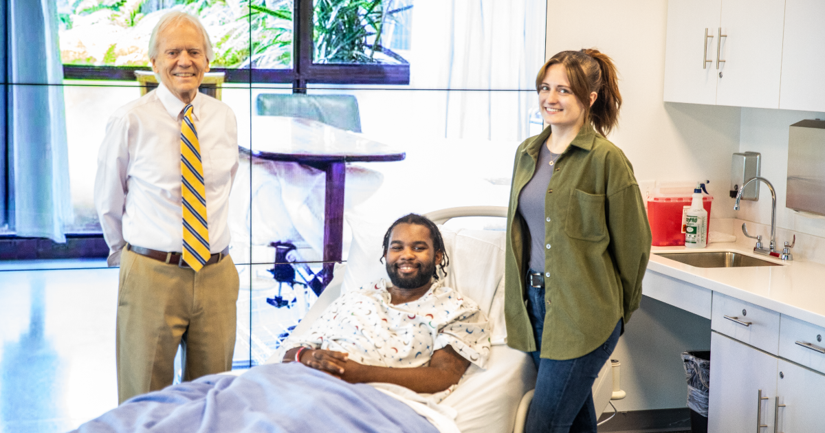 A man and woman stand around a man posing as a patient in a hospital bed 
