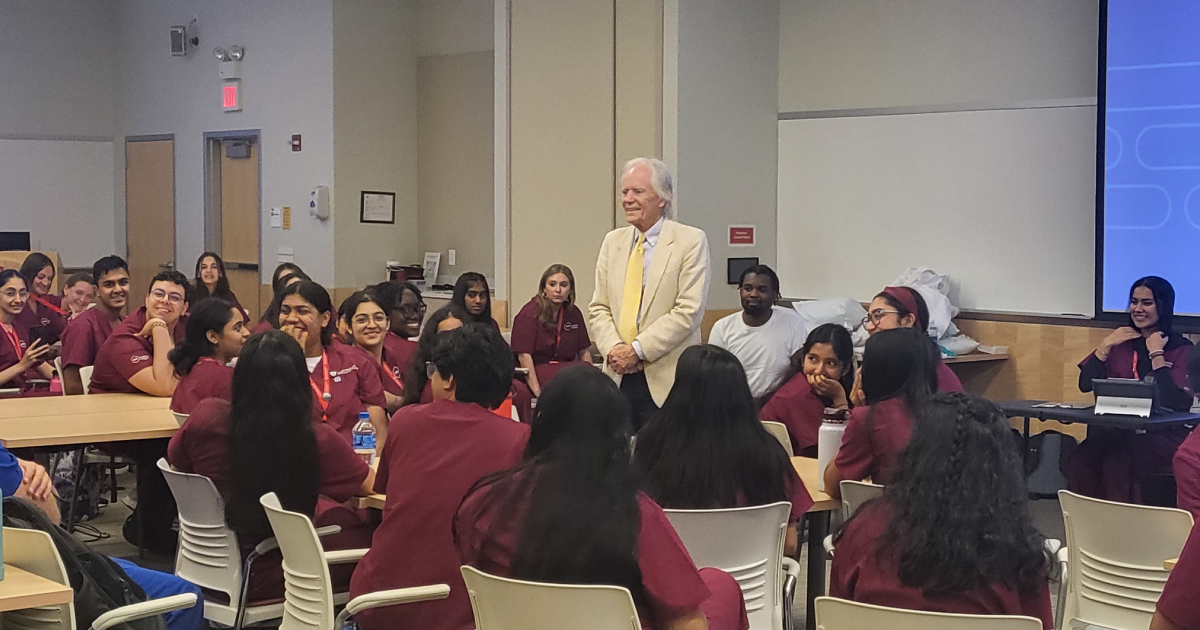 A man stands speaking and surrounded in a class full of healthcare students 