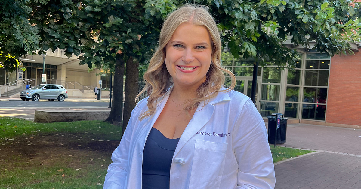 University of Delaware College of Health Sciences alumna Margaret Downing dons her white PA coat posing for a photo outdoors.. 