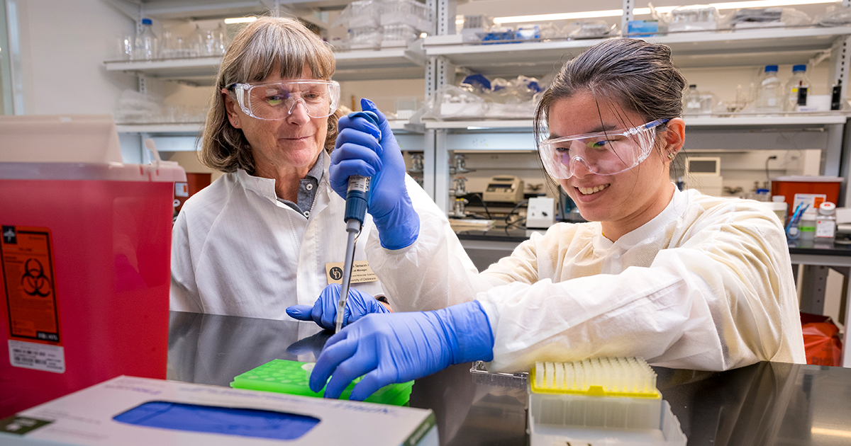 A student and professor work in a lab wearing goggles and blue latex gloves holding a pipette.