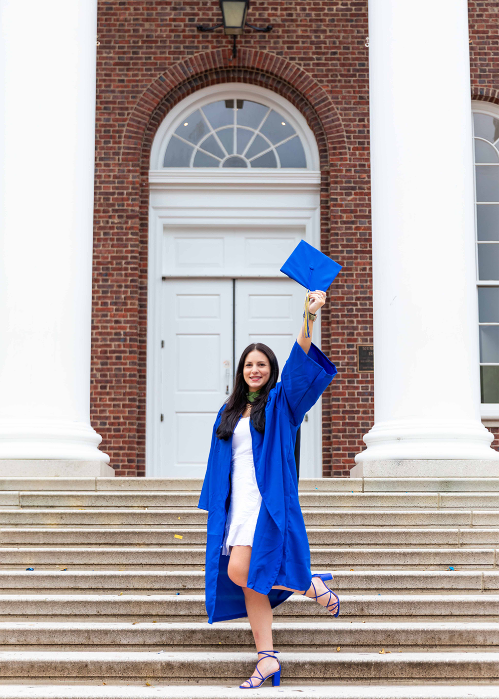 Senior Erika Scholtz in her blue UD graduation regalia holds her diploma over her head in front posing on the stairs of Memorial Hall.