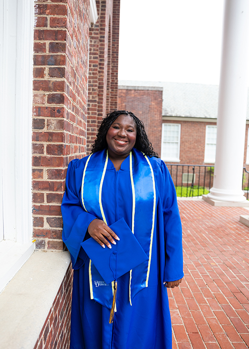 Ciara Mbuakoto wears her UD Commencement regalia holding her diploma in her hand as she poses outside Memorial Hall