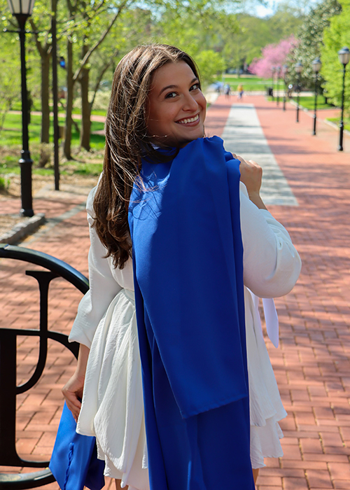 Shayna DeRosa poses on UD's campus with her UD Commencement gown slung over her shoulder as she turns back and looks at the camera.