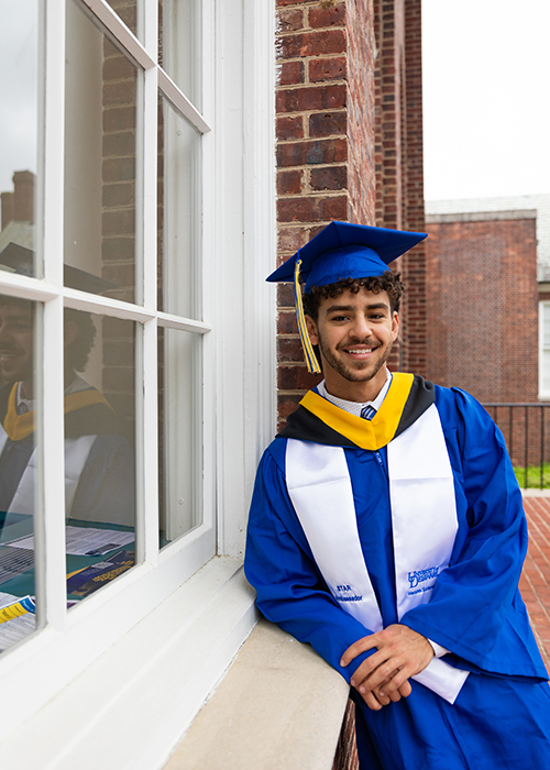 Menelik Duey, a senior applied molecular biology and biotechnology major, Menelik Duey, a senior applied molecular biology and biotechnology major, poses wearing his UD cap and gown while leaning on a window of Memorial Hall.