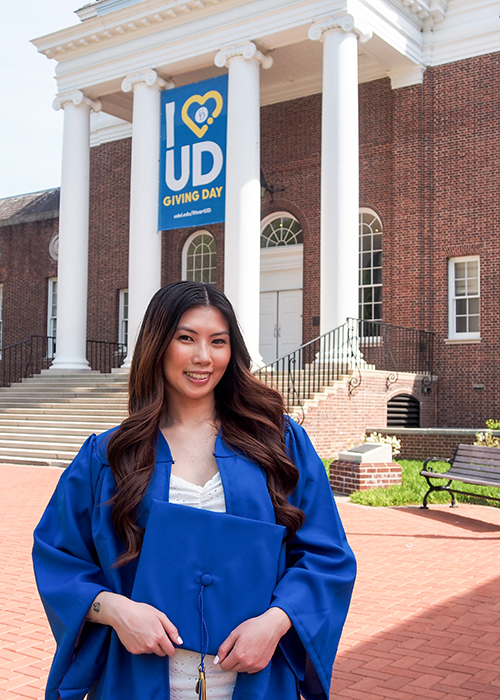Emily Chan, a medical laboratory science major, wears her graduation regalia, holding her cap, and poses outside Memorial Hall where an I Love UD banner hangs behind her.
