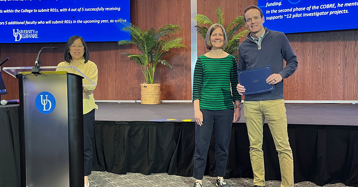 Dave Edwards, Chair of the Department of Kinesiology and Applied Physiology poses with Jocelyn Hafer, Assistant KAAP Professor who's on the awards committee. Edwards won the Excellence in Mentorship of Faculty Award. On the far left, CHS Awards Committee Chair Shea Ching Chai stands at a UD podium, announcing winners. 