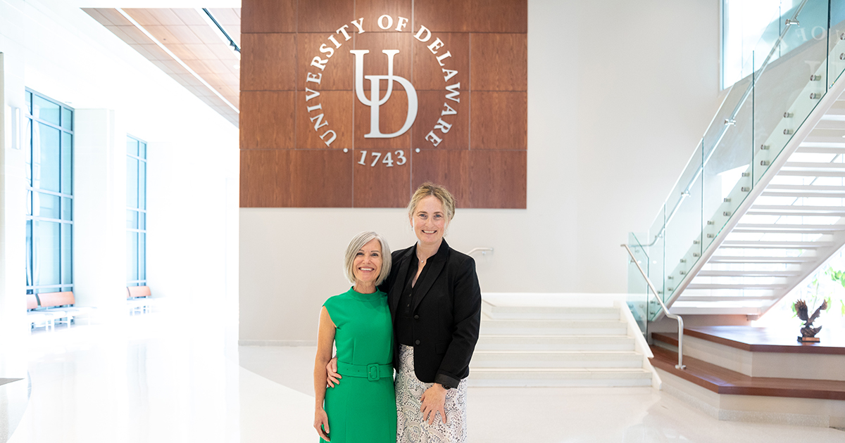 Winston Churchill Fellow Heather McFarlane of Northern Ireland wears a green dress on the left and poses with Freda Patterson, associate dean of research for CHS, with the UD logo behind them in the lobby of STAR Tower.